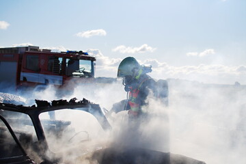 A firefighter with a breathing apparatus extinguishes a car fire against the rays of the sun with smoke and water splinters in the background