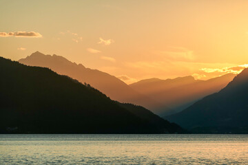 A sunset by Millstaetter lake in Austria. The lake is surrounded by high Alps. Calm surface of the lake reflecting the sunbeams. The sun sets behind the mountains. A bit of overcast. Natural beauty
