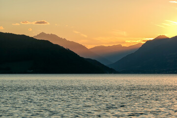 A sunset by Millstaetter lake in Austria. The lake is surrounded by high Alps. Calm surface of the lake reflecting the sunbeams. The sun sets behind the mountains. A bit of overcast. Natural beauty