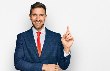 Handsome man with beard wearing business suit and tie with a big smile on face, pointing with hand and finger to the side looking at the camera.