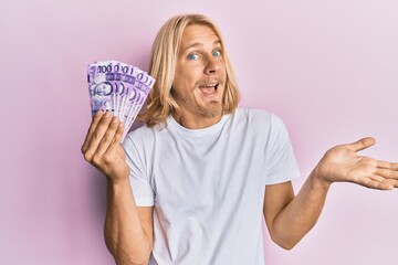 Caucasian young man with long hair holding 100 philippine peso banknotes celebrating achievement with happy smile and winner expression with raised hand