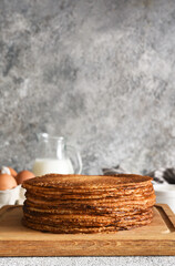 stack of thin pancakes on a wooden board. Pancake ingredients: flour, milk, eggs, whisk on the kitchen table.