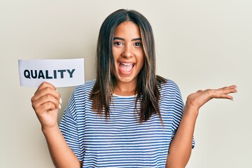 Young latin girl holding quality word on paper celebrating victory with happy smile and winner expression with raised hands