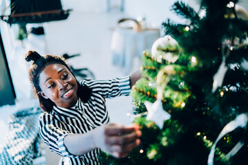 Happy young African American woman decorating fir tree at home