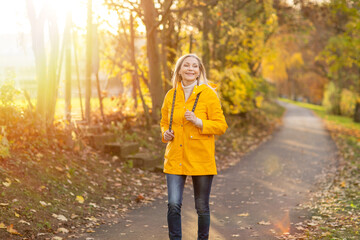 Weißhaarige Grauhaarige Frau spaziert im Park Herbst Sonnenstrahlen gelbe Regenmantel  - obrazy, fototapety, plakaty