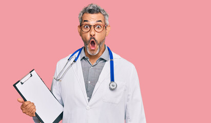 Middle age grey-haired man wearing doctor stethoscope holding clipboard scared and amazed with open mouth for surprise, disbelief face