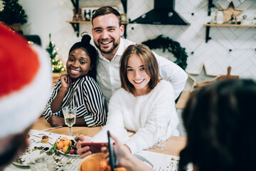 Cheerful gathering of young women and men browsing mobile while celebrating Christmas at home
