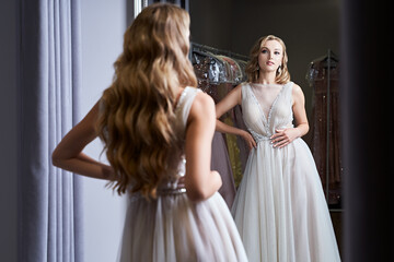 Fototapeta na wymiar Young beautiful blonde girl wearing a full-length silver white chiffon prom ball gown decorated with sparkles and sequins. Model in front of mirror in a fitting room at dress hire service.