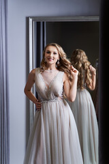 Young beautiful blonde girl wearing a full-length silver white chiffon prom ball gown decorated with sparkles and sequins. Model in front of mirror in a fitting room at dress hire service.