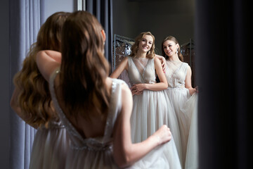 Two young beautiful girls wearing a full-length silver white chiffon prom ball gowns decorated with sparkles and sequins. Models in front of mirror in a fitting room.