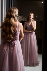 Young beautiful blonde girl wearing a full-length pink violet or purple chiffon prom ball gown decorated with sparkles and sequins. Model in front of mirror in a fitting room at dress hire service.