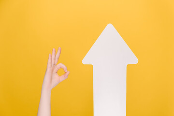 Naklejka premium Image of unrecognizable female makes thumb up gesture near white paper arrow, demonstrates approval or agreement, isolated on yellow background. Body language concept. Hand sign. Like gesture