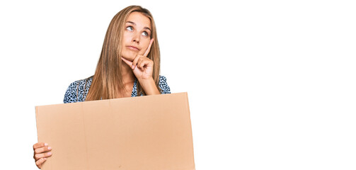 Young blonde woman holding cardboard empty banner serious face thinking about question with hand on...