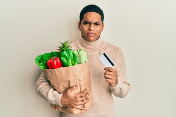 Young handsome hispanic man holding groceries and credit card skeptic and nervous, frowning upset because of problem. negative person.