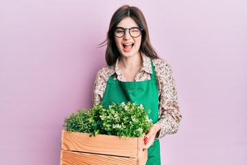 Young beautiful caucasian girl wearing gardener apron holding wooden plant pot winking looking at the camera with sexy expression, cheerful and happy face.