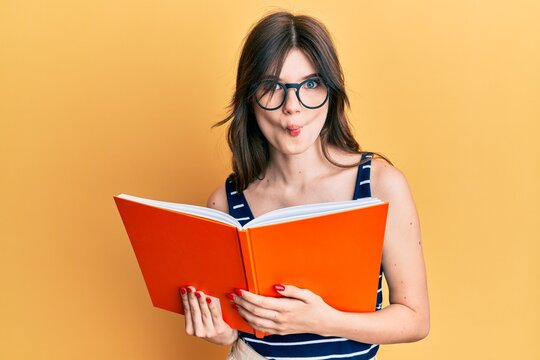 Young beautiful caucasian girl reading a book wearing glasses making fish face with mouth and squinting eyes, crazy and comical.