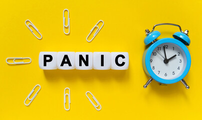 On a yellow background, a blue alarm clock, paper clips and white cubes on which the text is written - PANIC
