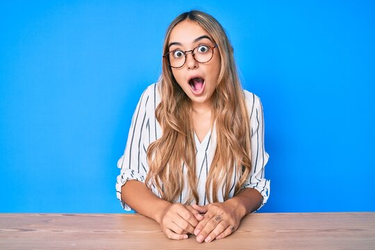 Young beautiful blonde woman wearing glasses sitting on the table scared and amazed with open mouth for surprise, disbelief face