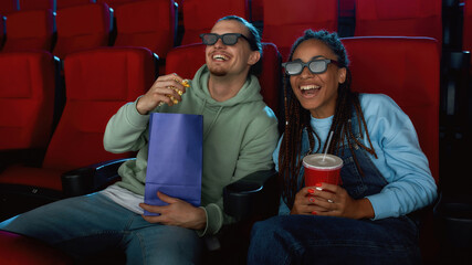 Cheerful young couple wearing 3d glasses laughing, while watching a movie together in cinema auditorium, eating popcorn and drinking soda