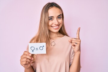 Young beautiful blonde woman holding we are equal paper surprised with an idea or question pointing finger with happy face, number one