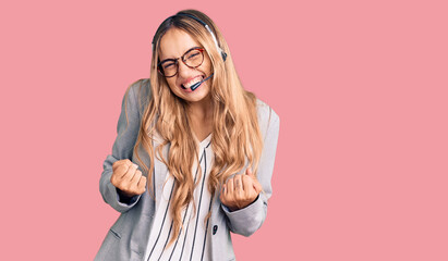Young beautiful blonde woman wearing call center agent headset very happy and excited doing winner...
