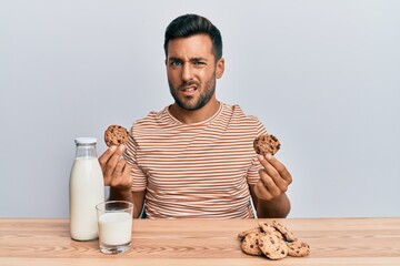 Handsome hispanic man drinking a glass of milk holding chocolate cookies clueless and confused...