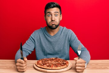 Handsome hispanic man eating tasty pepperoni pizza puffing cheeks with funny face. mouth inflated with air, catching air.