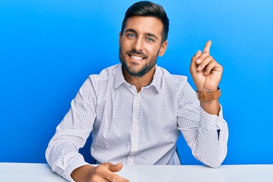 Handsome hispanic man wearing business clothes sitting on the table with a big smile on face, pointing with hand finger to the side looking at the camera.