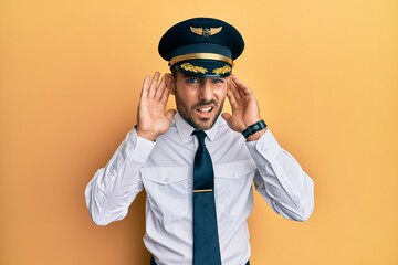 Handsome hispanic man wearing airplane pilot uniform trying to hear both hands on ear gesture, curious for gossip. hearing problem, deaf © Krakenimages.com
