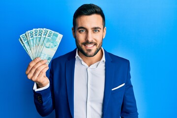 Young hispanic businessman wearing business suit holding brazilian real banknotes looking positive...