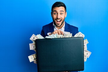 Young hispanic businessman holding briefcase full of dollars smiling and laughing hard out loud...