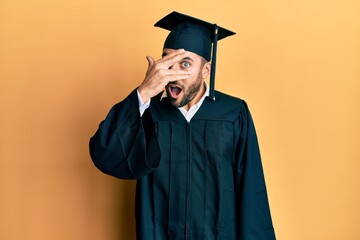 Young hispanic man wearing graduation cap and ceremony robe peeking in shock covering face and eyes with hand, looking through fingers with embarrassed expression.