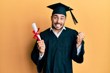 Young hispanic man wearing graduation robe holding diploma screaming proud, celebrating victory and...