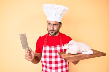 Young hispanic man cooking chicken holding knife skeptic and nervous, frowning upset because of...
