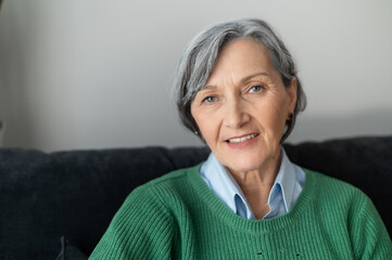 A headshot of a charming beaming old lady, gray-haired senior mature woman sitting in the living room and looking at the camera, wearing a casual green jumper, resting and smiling