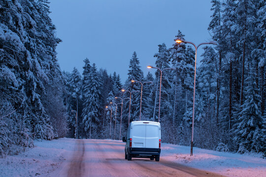 A white cargo delivery courier van driving in the snowy winter suburb road in the evening with trees on roadside and streetlights illuminated