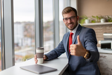 Fototapeta premium Smiling businessman having a coffee break at the office near a window and showing thumb up.