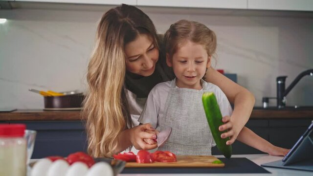 close up, camera dolly tracking, moving background: young mother smiles and kisses her young daughter with long hair, they stand in kitchen, and child trains to scatter exactly tomato round slices