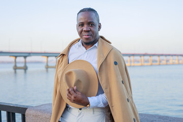 Stylish African- American man in a hat, shirt and trousers on the background of the river