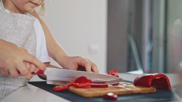 camera left tracking: mom holds her daughter's hand in her hand, cuts together red Bulgarian fresh pepper, girl learns to cook salads