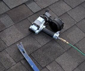 Two tools used by a professional roofer: pry bar to loosen nails and remove damaged shingles; nailg un to secure new shingles in place.