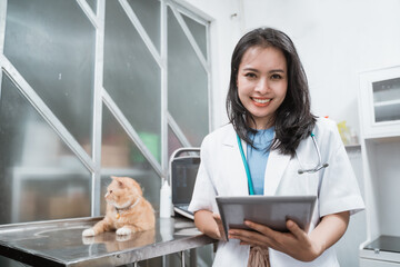 young veterinarian smiling at camera while using digital tablet near a cat sits on the table at the vet clinic