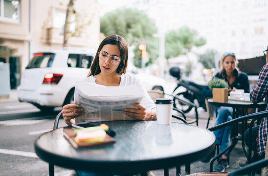 Serious caucasian woman in eyewear for vision correction sitting at table on cfe terrace reading morning news in daily printed media, pensive female reading holding newspaper searching vacancy