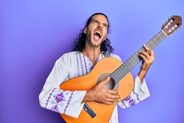 Fototapeta premium Young handsome man with long hair playing classical guitar angry and mad screaming frustrated and furious, shouting with anger looking up.