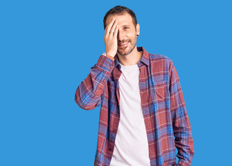 Young handsome man wearing casual clothes covering one eye with hand, confident smile on face and surprise emotion.