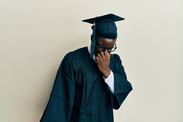 Handsome black man wearing graduation cap and ceremony robe smelling something stinky and disgusting, intolerable smell, holding breath with fingers on nose. bad smell