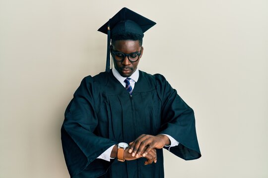 Handsome black man wearing graduation cap and ceremony robe checking the time on wrist watch, relaxed and confident