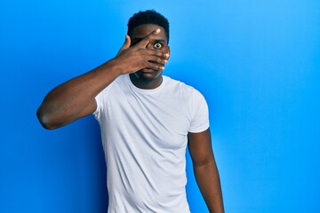 Handsome black man wearing casual white t shirt peeking in shock covering face and eyes with hand, looking through fingers afraid