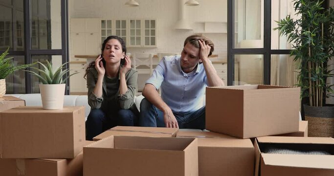 Unhappy young family couple feeling stressed moving into new apartment or moving out of house, sitting together on sofa among huge carton boxes, tired of long renovation relocation processes.