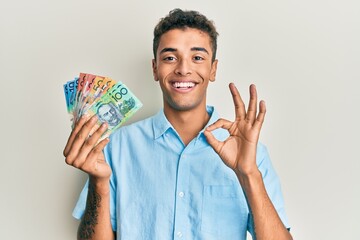 Young handsome african american man holding australian dollars doing ok sign with fingers, smiling friendly gesturing excellent symbol